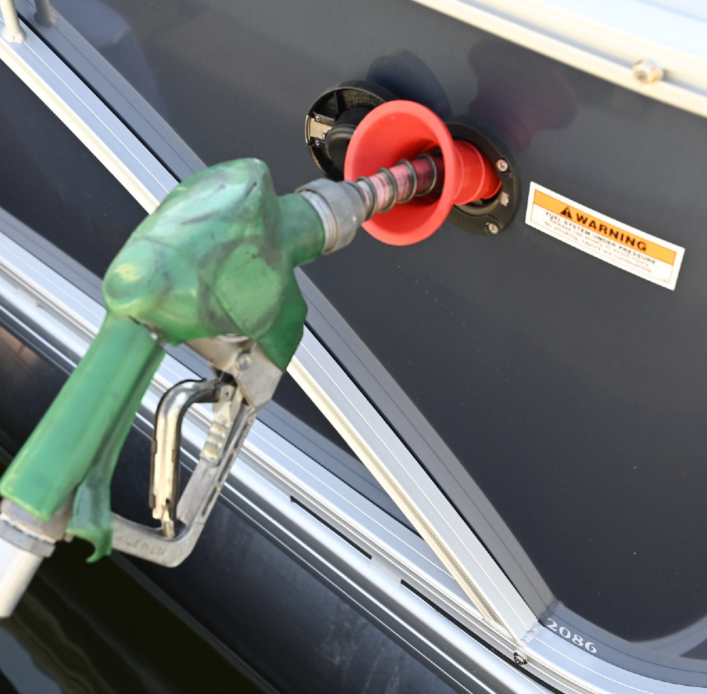 A green fuel nozzle pumping gas into a pontoon boat's fuel port using a red plastic funnel adapter.