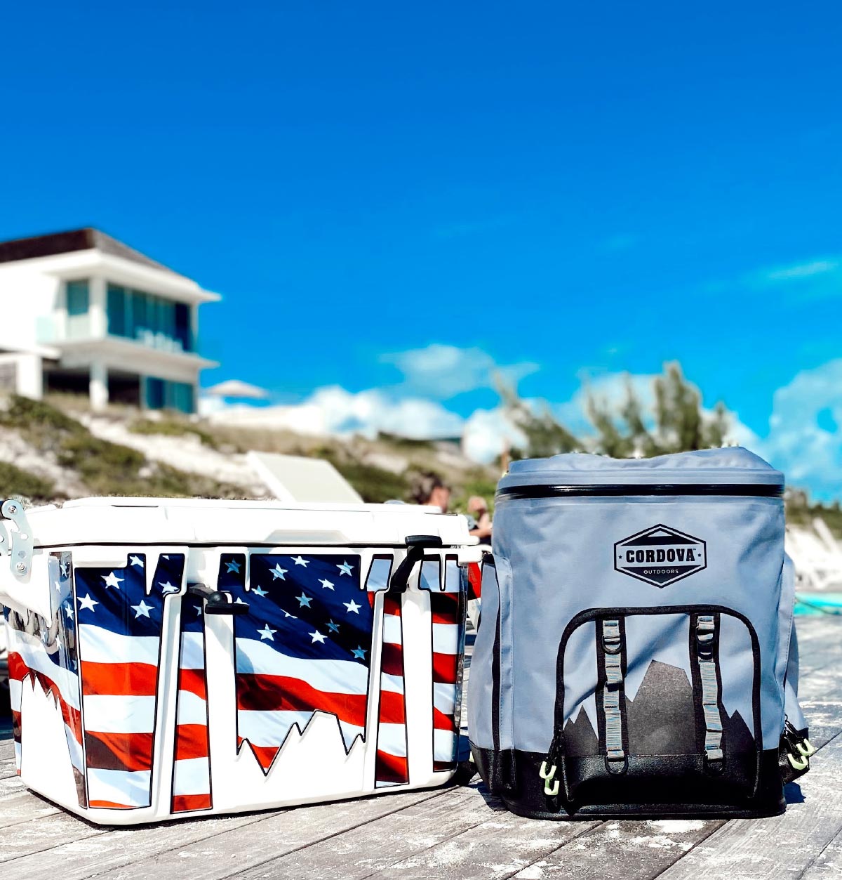 An American flag themed hard cooler sits next to a grey Cordova soft-sided cooler on a sunny wooden dock.