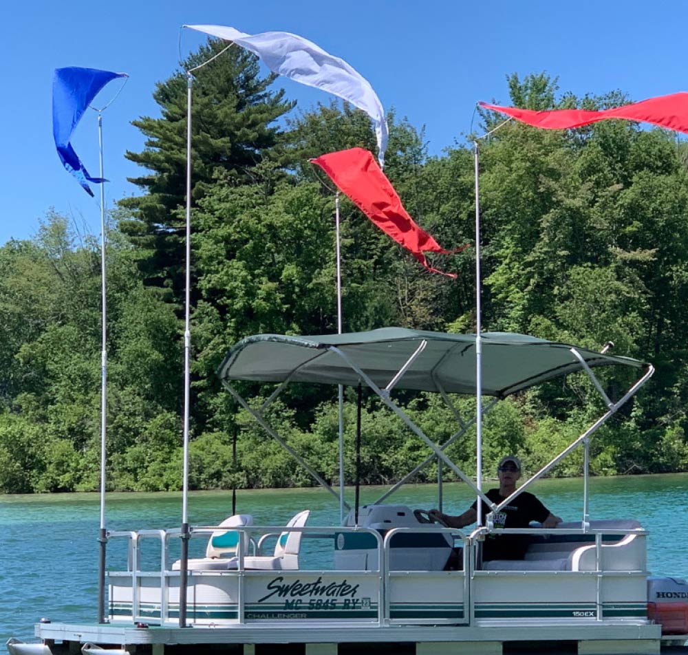 A Sweetwater pontoon boat on a lake flying tall, colorful blue, white, and red wind flags.