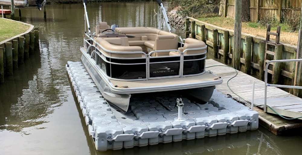 A tan Bennington pontoon boat resting on a grey modular floating drive-on boat lift next to a dock.