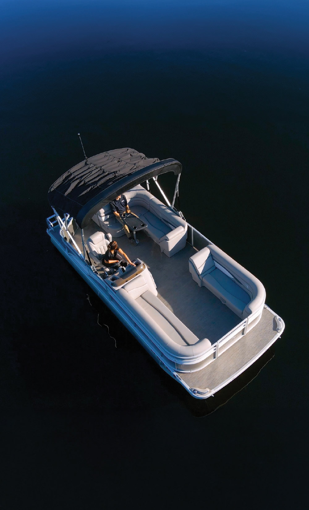 High-angle aerial view of a grey pontoon boat with a black bimini top drifting on calm, dark water.