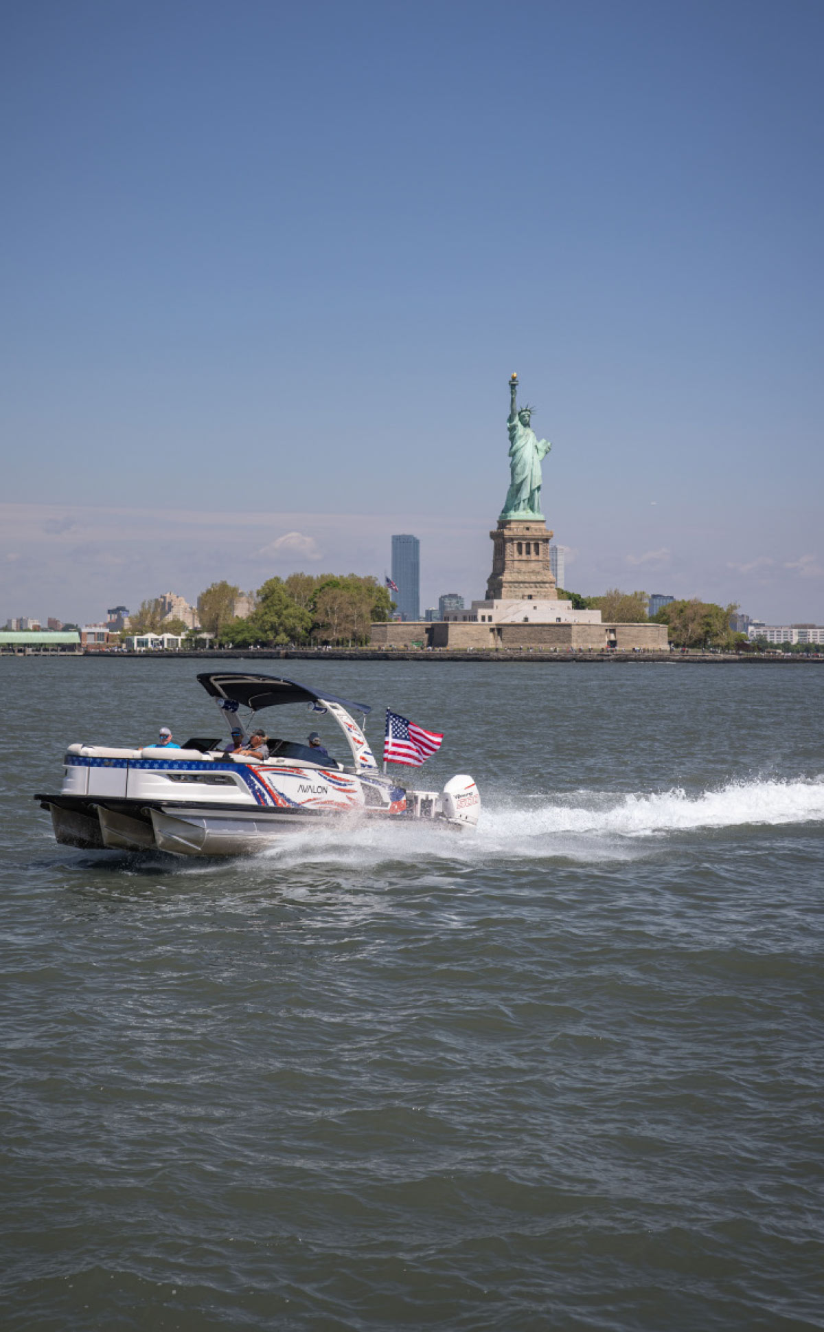 An Avalon pontoon boat with a U.S. flag speeds across the water, with the Statue of Liberty standing prominently in the background.