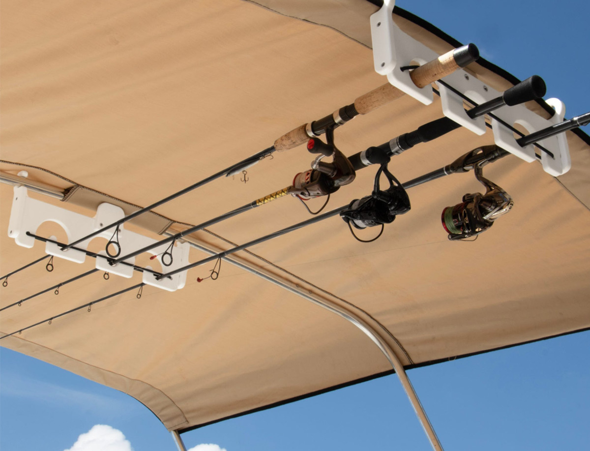 Three fishing rods are stored horizontally under a tan boat bimini top canopy using white Boat Outfitters rod holders. The rods and reels are securely mounted to the overhead frame against a clear blue sky.