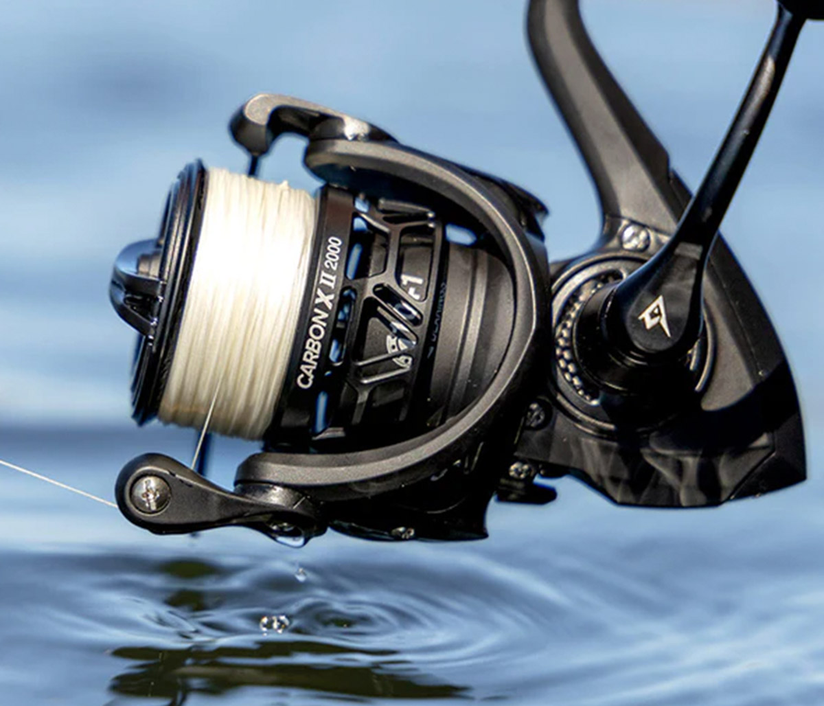 A close-up of a black Piscifun Carbon X II 2000 spinning reel spooled with white fishing line. The reel is positioned just above a calm, rippling water surface, with a single water droplet dripping from its lower edge.