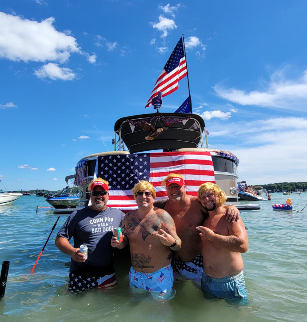 Four men wearing blonde wigs stand waist-deep in water in front of a pontoon boat. The boat is decorated with a large American flag. One man wears a shirt reading "CORN POP WAS A BAD DUDE," while the others are shirtless.