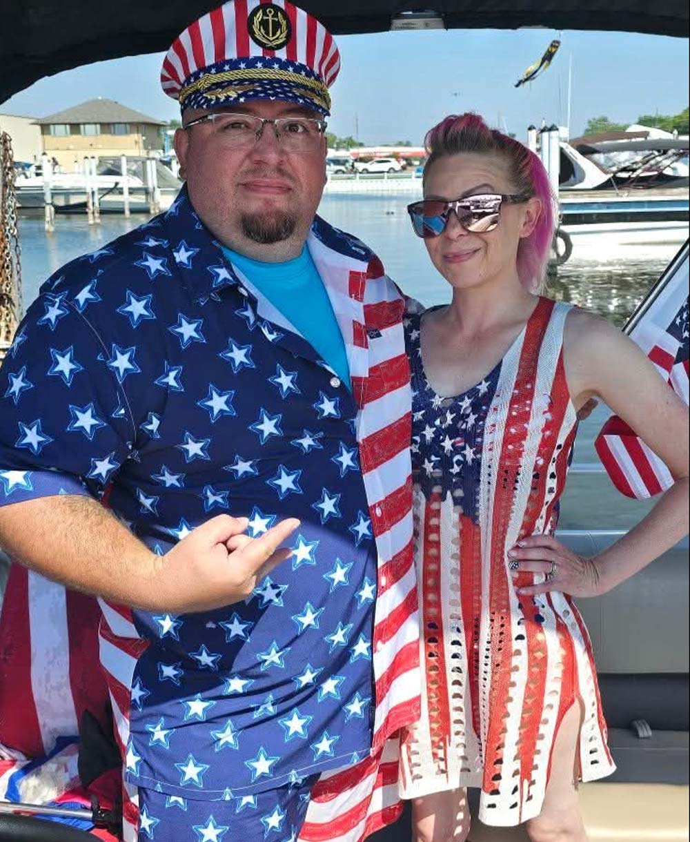 A man wearing glasses, a patriotic captain's hat, and a star-patterned shirt stands next to a woman with pink hair and sunglasses wearing a red, white, and blue patterned dress on a boat.