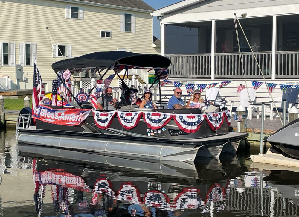 Four people sit on a pontoon boat docked near waterfront homes. The boat is heavily decorated with American flags, patriotic pinwheels, and red, white, and blue bunting banners, reflecting in the calm water.