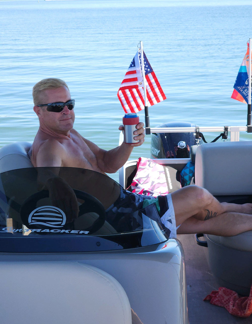 A shirtless man wearing sunglasses relaxes on a Sun Tracker pontoon boat, holding up a beverage can in a red and blue koozie. An American flag flies at the stern over calm blue water.