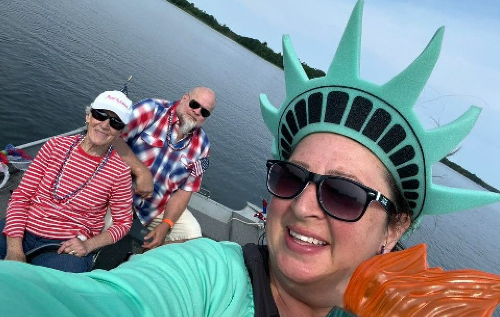 A woman takes a selfie wearing sunglasses, a green foam Statue of Liberty crown, and holding a plastic torch. In the background, two people sit on a boat on open water.
