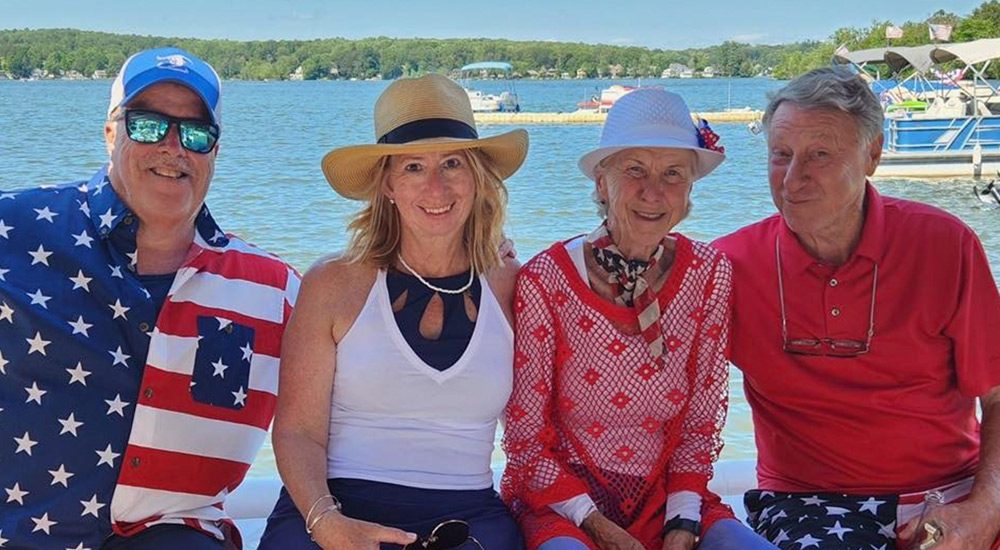 Four people sit side-by-side on a boat with water and a distant shoreline behind them. They wear summer clothes and hats, including a shirt with an American flag pattern and a patriotic neck scarf.