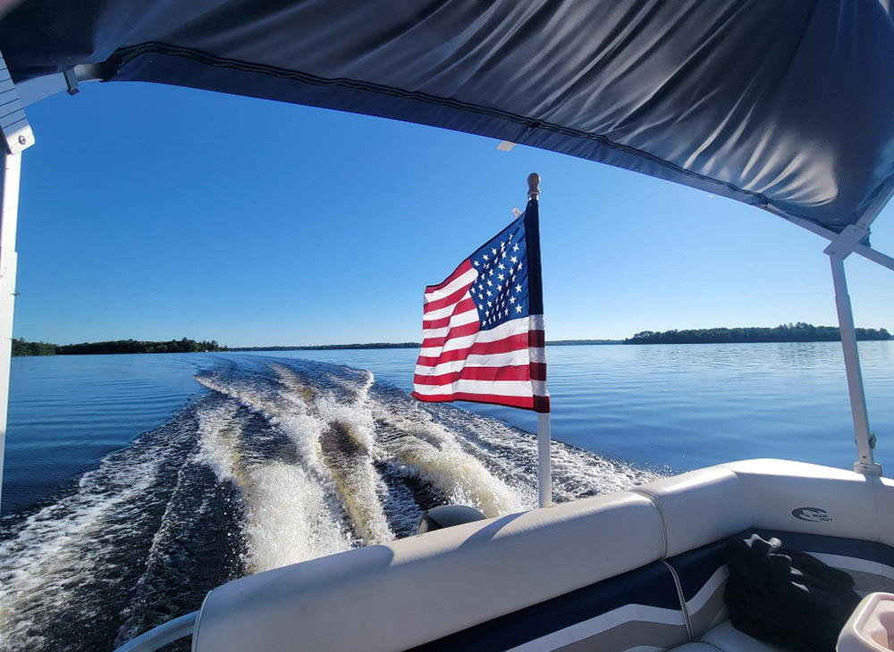 View from the back of a boat under a canopy looking out at open blue water. An American flag flies from a stern pole over a wide white wake stretching toward a clear blue sky and distant land.