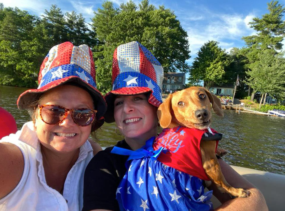Two women wearing red, white, and blue sequined top hats pose on a boat with a small brown dog in a patriotic star-patterned outfit. A tree-lined shore and water are visible in the background.