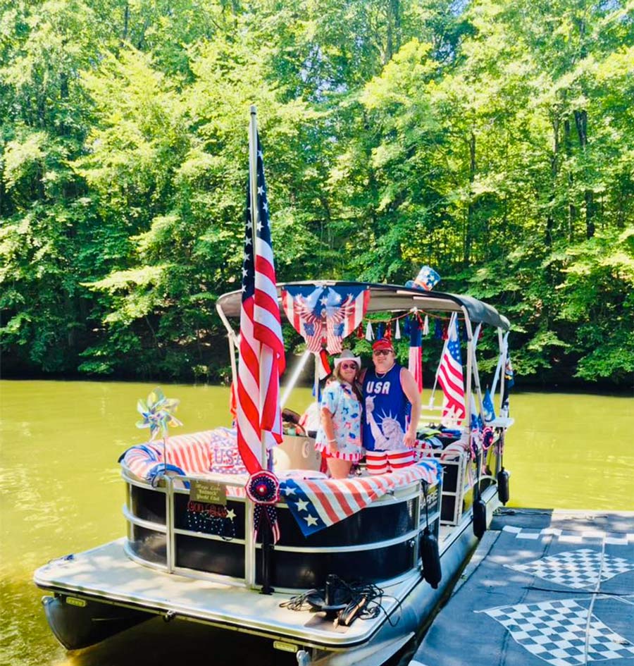 A man and woman stand on a pontoon boat heavily decorated with American flags, patriotic banners, and red, white, and blue bunting. The boat is docked near a dense green forest lining a calm river.