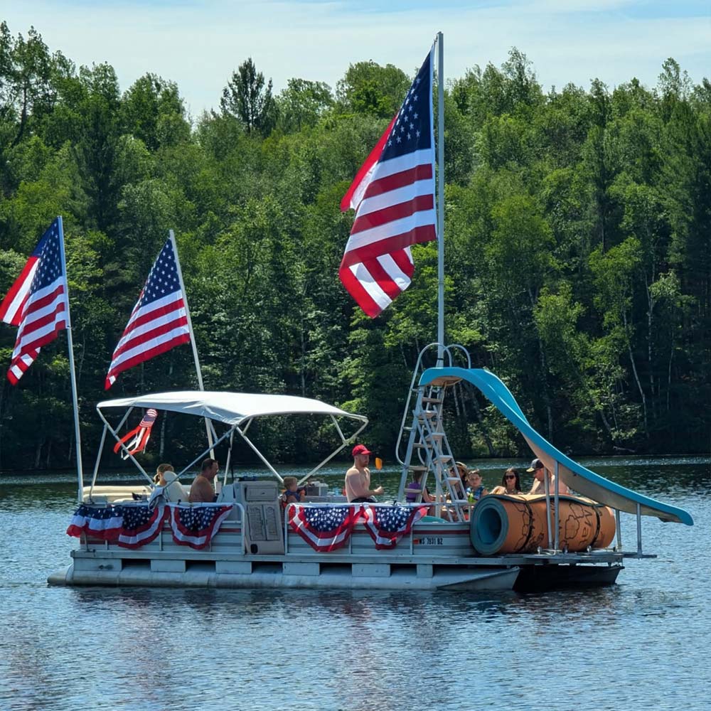 A pontoon boat carrying passengers floats on a lake. The boat features a built-in water slide, red, white, and blue patriotic bunting along the railings, and three large American flags flying from tall stern poles.