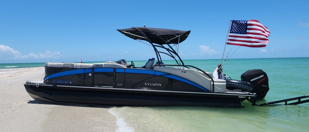 A black and grey Sylvan pontoon boat with blue trim is parked on a sandy beach where clear turquoise water meets the shore. An American flag flies from the stern near a black Mercury outboard motor under a bright blue sky.