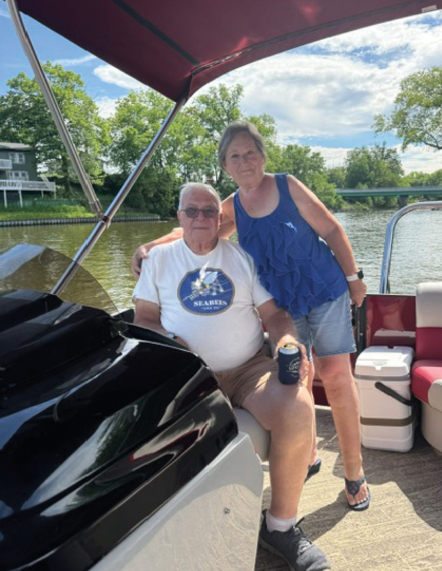 An elderly couple poses together smiling on a pontoon boat on the water.