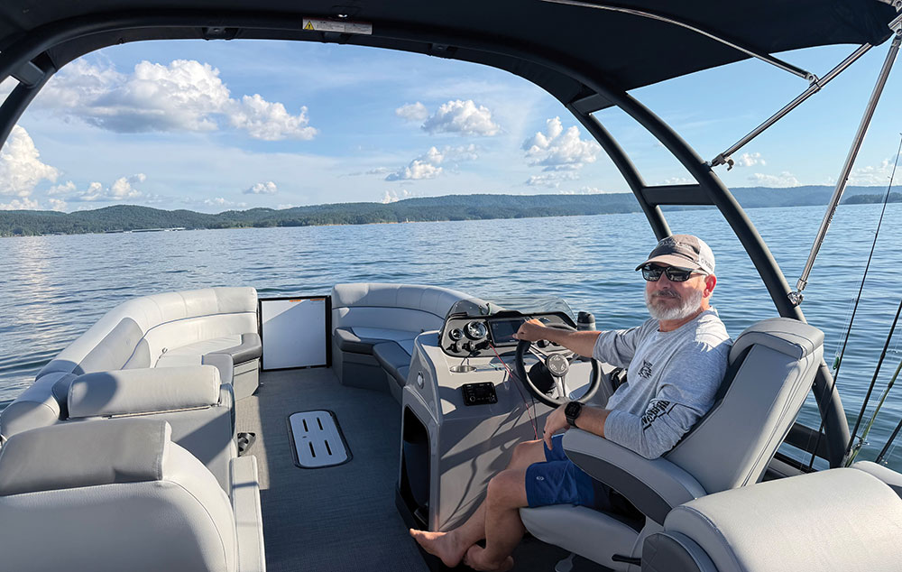 A man sitting at the steering helm of a pontoon boat on a lake.