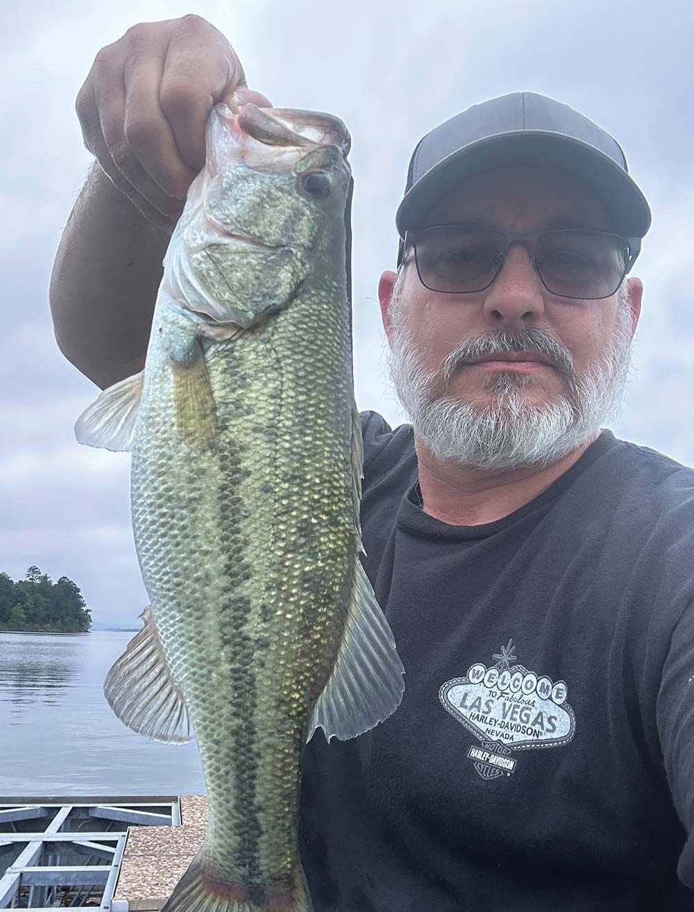 A close-up selfie of a man holding a large bass he caught on a lake.