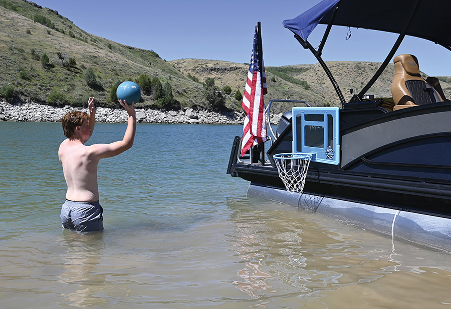 A boy stands in the water holding a basketball near a boat.