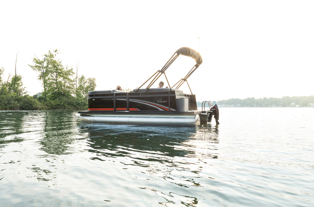 A Qwest pontoon boat floating on a quiet lake surrounded by trees during sunset.
