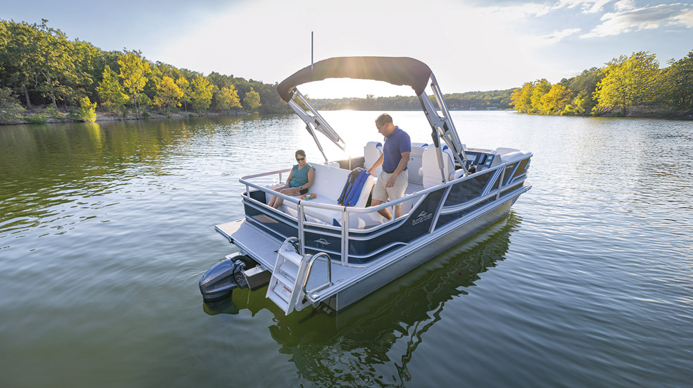 A man and woman relaxing on a SunCatcher pontoon boat anchored on a lake surrounded by trees.