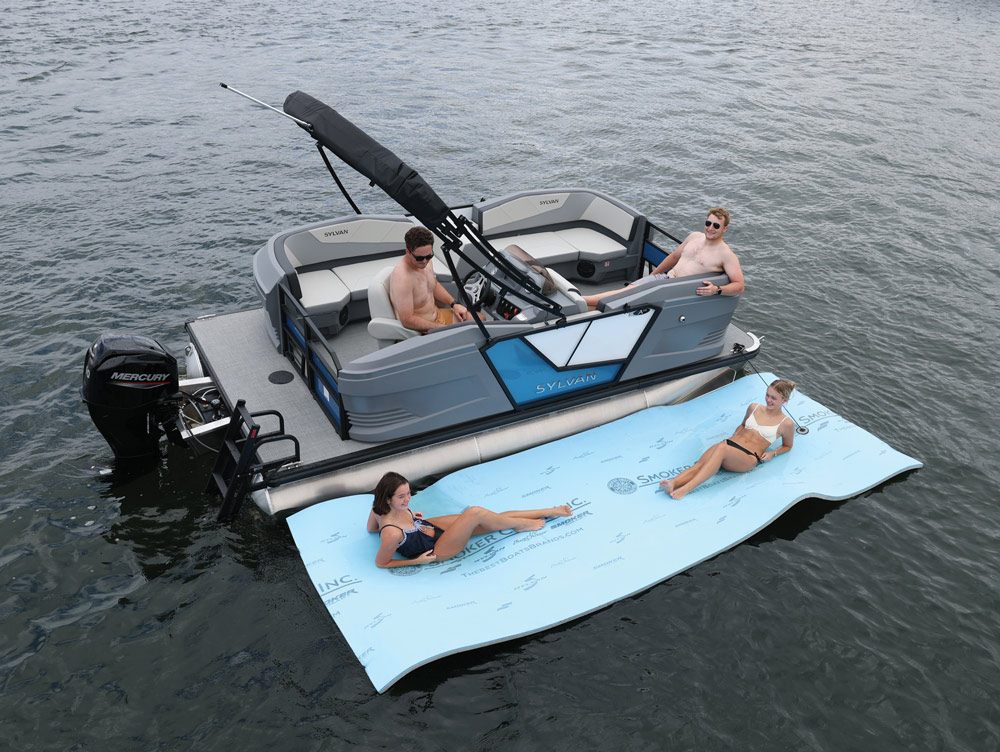 Two women relaxing on a floating mat tied to a Sylvan pontoon boat carrying two men.
