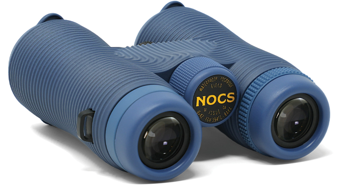A pair of blue binoculars with a distinct ribbed, textured grip housing, displayed on a white background. The central dial features the yellow brand name "NOCS".