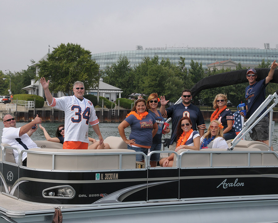 A group of sports fans in jerseys smiles and waves from a pontoon boat.