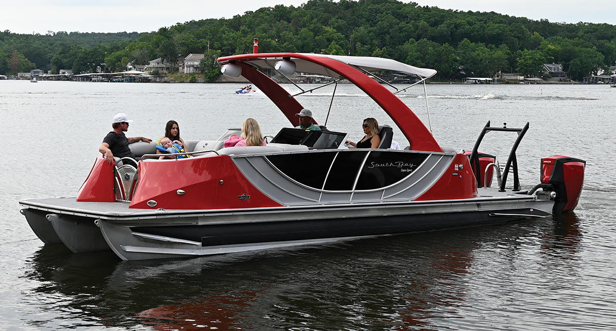 A red and grey South Bay pontoon boat carrying passengers glides across a lake.