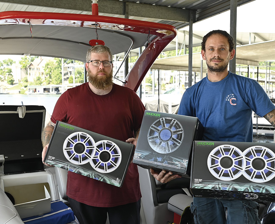 Two men pose inside a covered boat dock holding boxes of Kicker marine speakers.