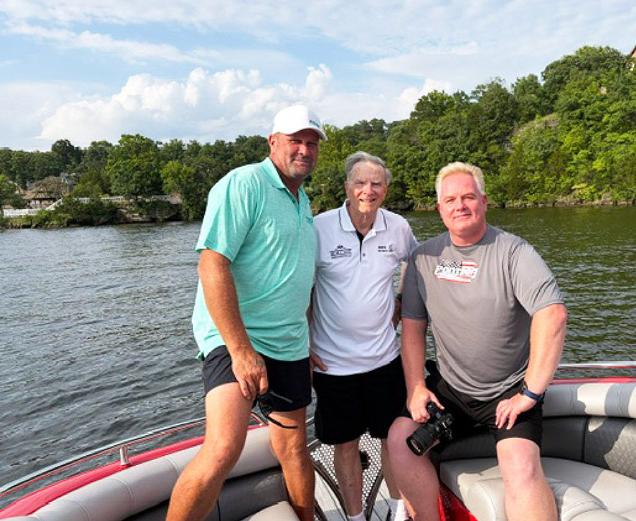 Three men pose together on a boat with trees and water in the background.