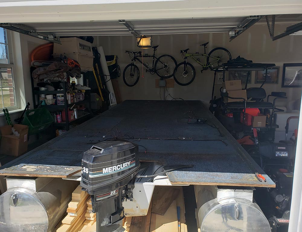A stripped-down pontoon boat deck and Mercury outboard motor inside a crowded garage.