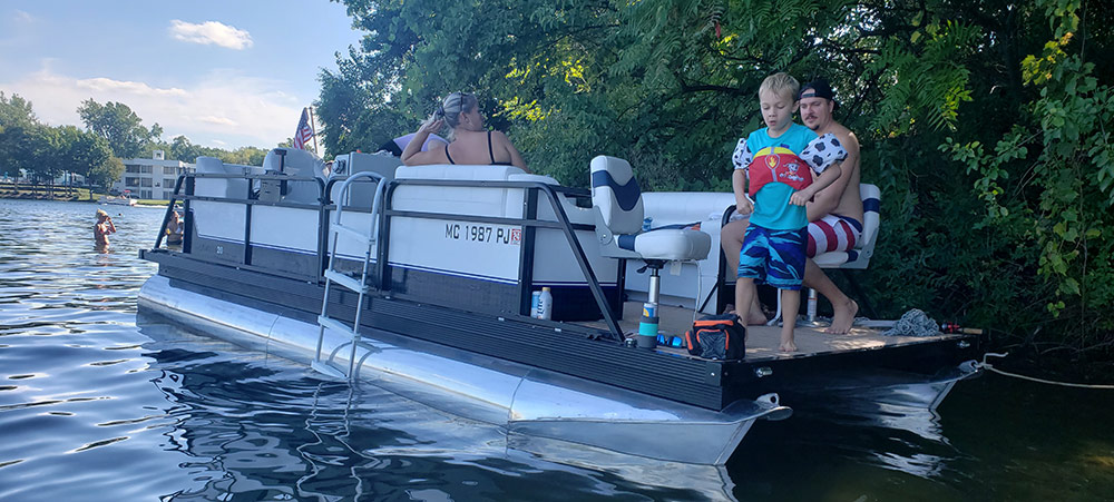 A family relaxing on their pontoon boat anchored near a shaded, tree-lined shore.