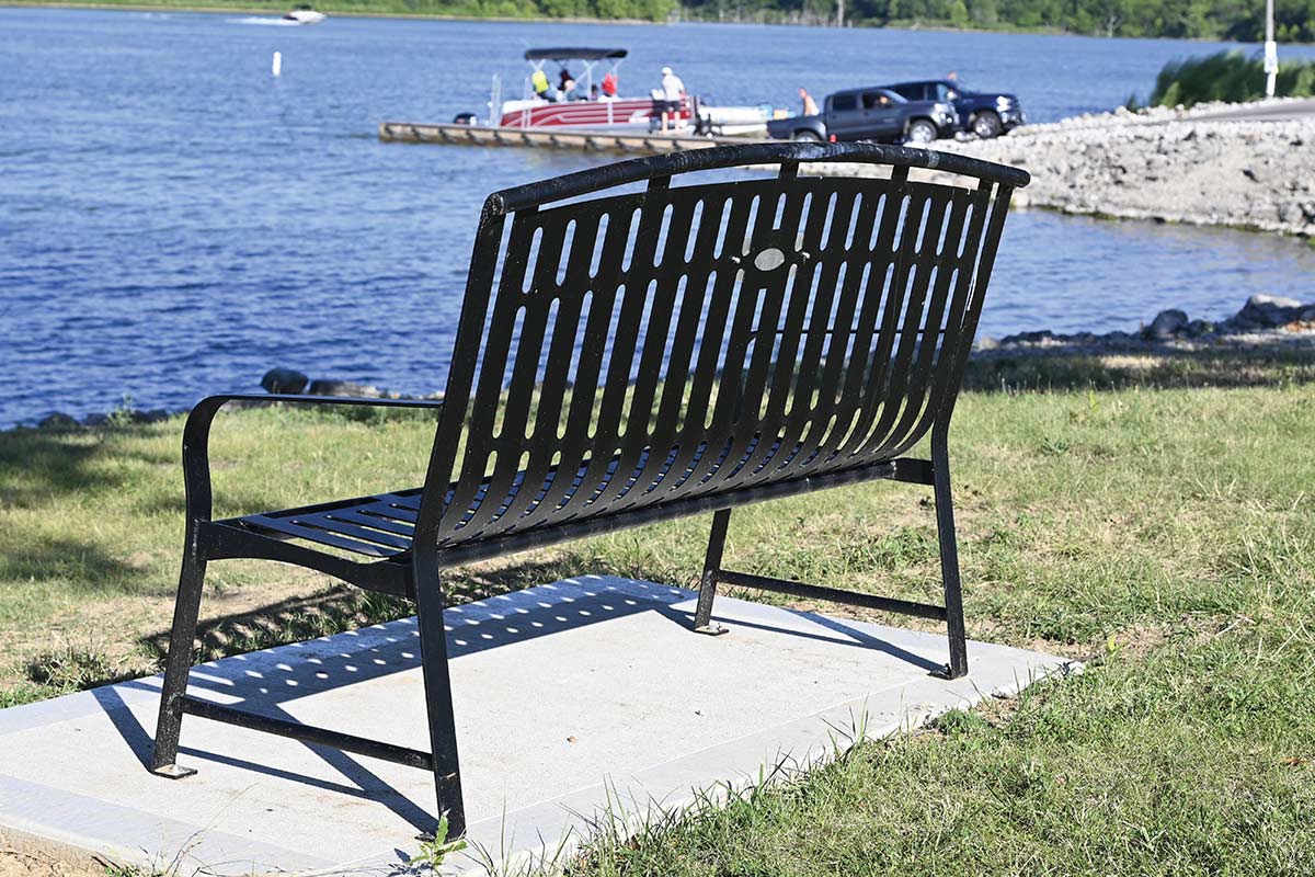 A black metal park bench facing a lake with a boat ramp in the background.