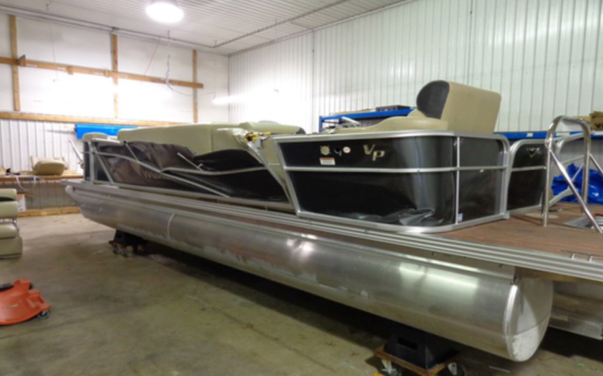 Manitou pontoon boat with damaged, crumpled side fencing resting inside a repair workshop.