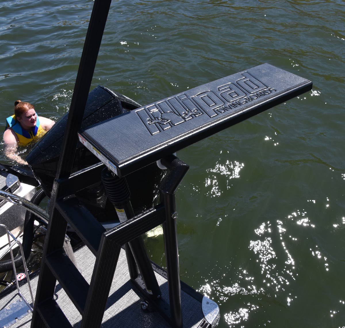 Black Lillipad diving board with a ladder attached to the rear deck of a pontoon boat on open water.