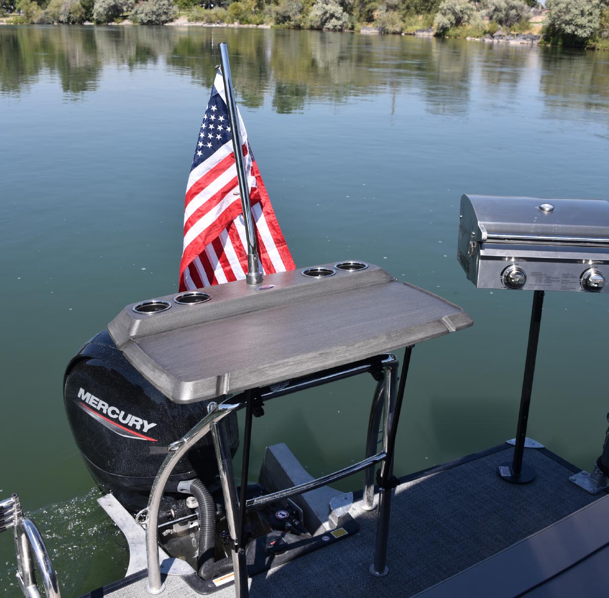 Boat stern featuring an American flag, a wood-grain prep table with cup holders, and a mounted stainless steel grill.