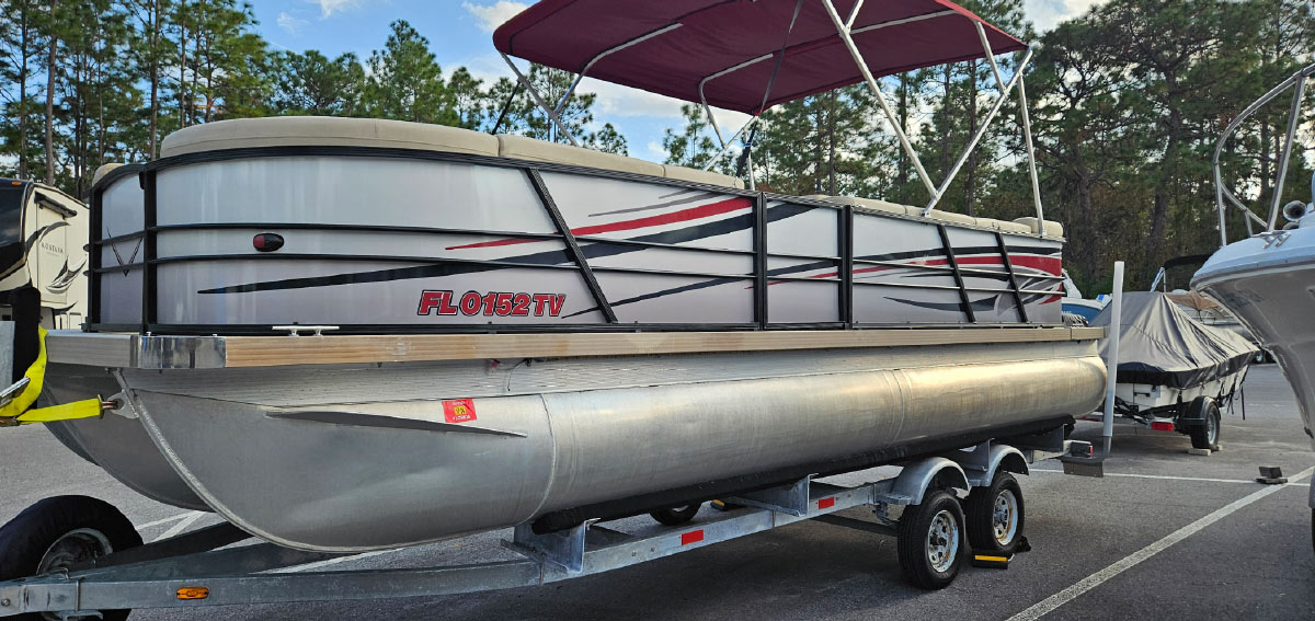 Silver pontoon boat with custom red-and-black graphic wraps and a maroon bimini top parked on a trailer.