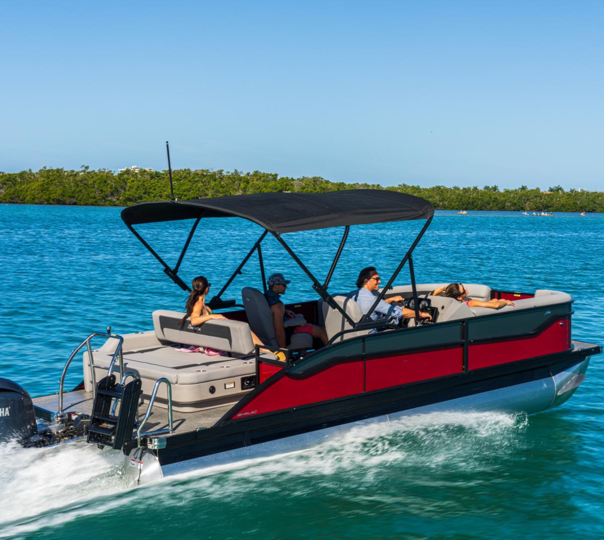 Red and grey Barletta pontoon boat cruising across bright blue water with passengers under a open black bimini top.