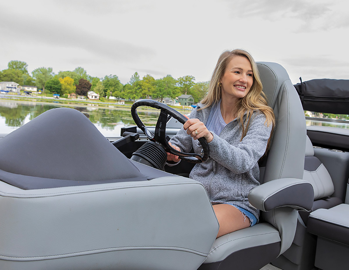 A smiling woman sitting in the captain's chair of a boat, holding the steering wheel while driving on a lake with a tree-lined shore in the background.