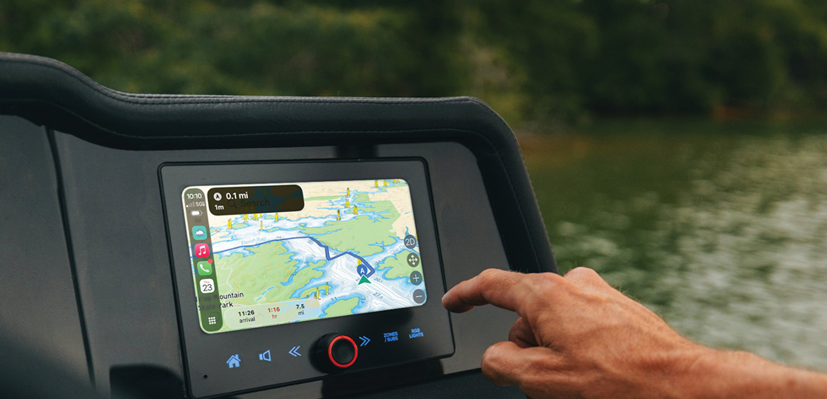 A hand touching a boat's touchscreen dashboard display, which shows Apple CarPlay GPS navigation with a waterway map while on the water.