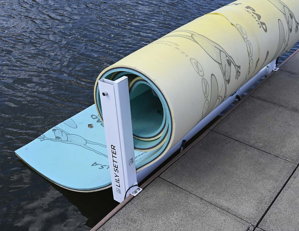 A rolled-up yellow and blue foam water mat stored in a white "Lily Setter" rack mounted on the edge of a boat dock over water.