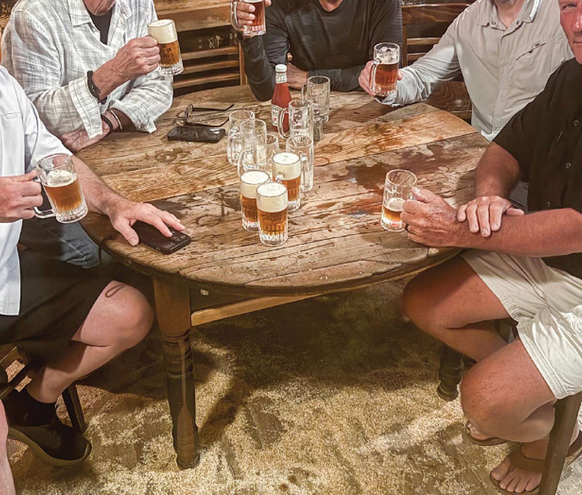 A group of men sitting around a round wooden table raising glass beer mugs for a toast, with several frothy beer glasses resting on the rustic table.