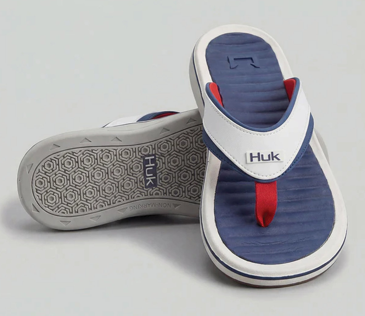 A pair of navy blue and white Huk flip-flops with red accents, displaying the cushioned footbed on one shoe and the grey non-marking treaded sole on the other.