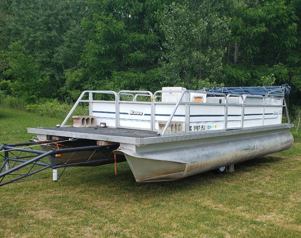 An older, weathered Raven pontoon boat sitting on a trailer in a grassy field.
