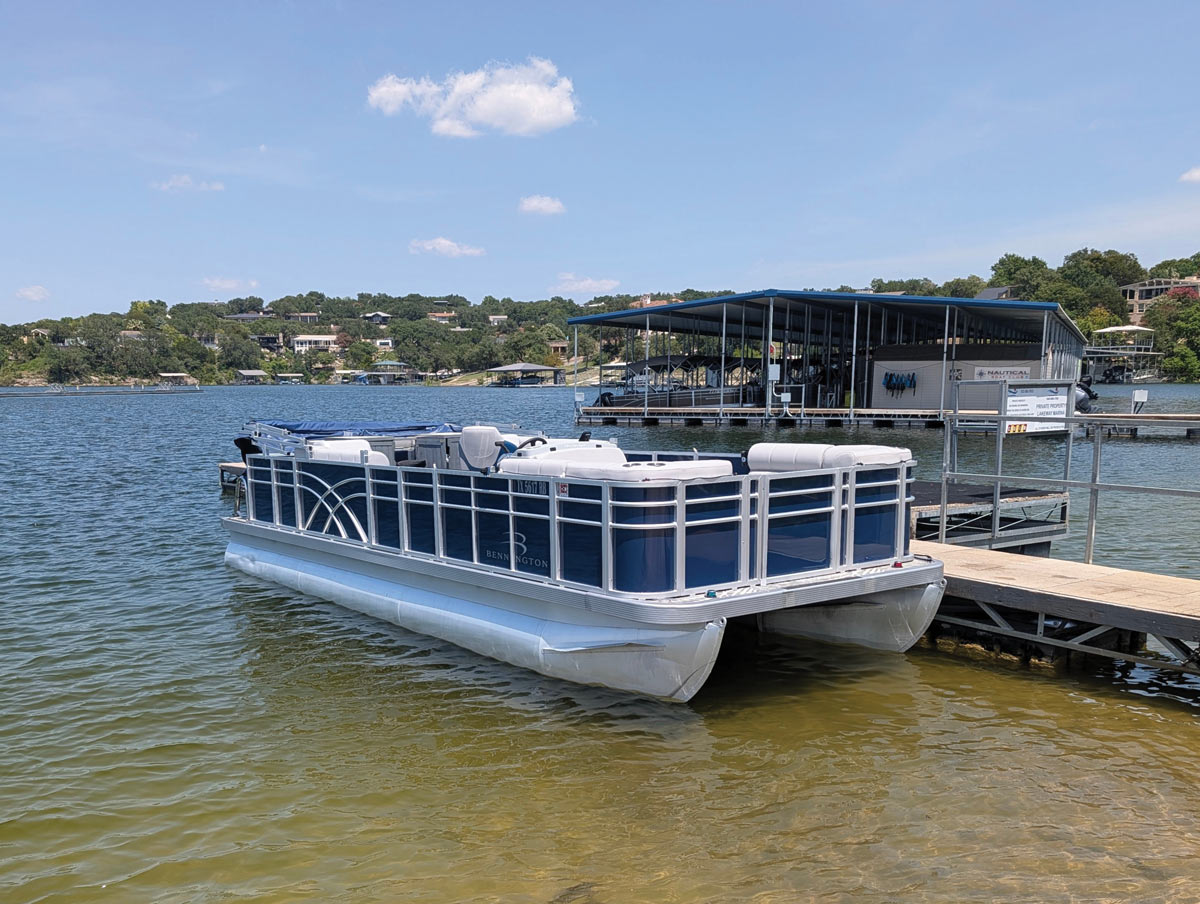 A blue and silver Bennington pontoon boat docked at a wooden pier on a sunny lake.