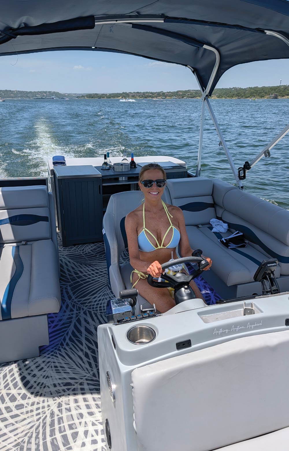 A woman in a bikini smiling while steering a pontoon boat with a bimini top on the water.