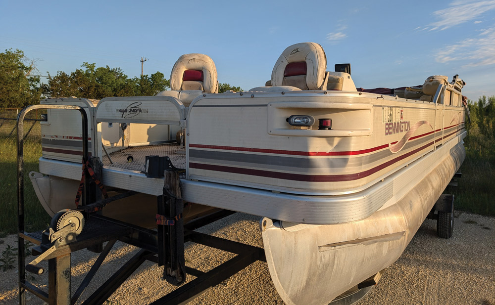 An old, weathered Bennington pontoon boat with faded red stripes sitting on a boat trailer.