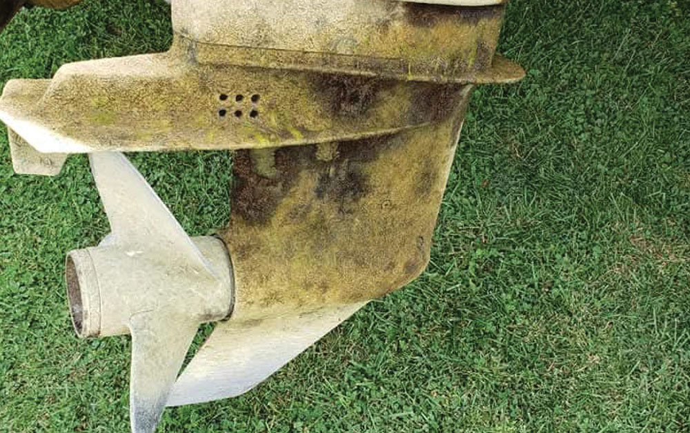 A grimy, algae-covered lower unit and propeller of an outboard motor resting on green grass.