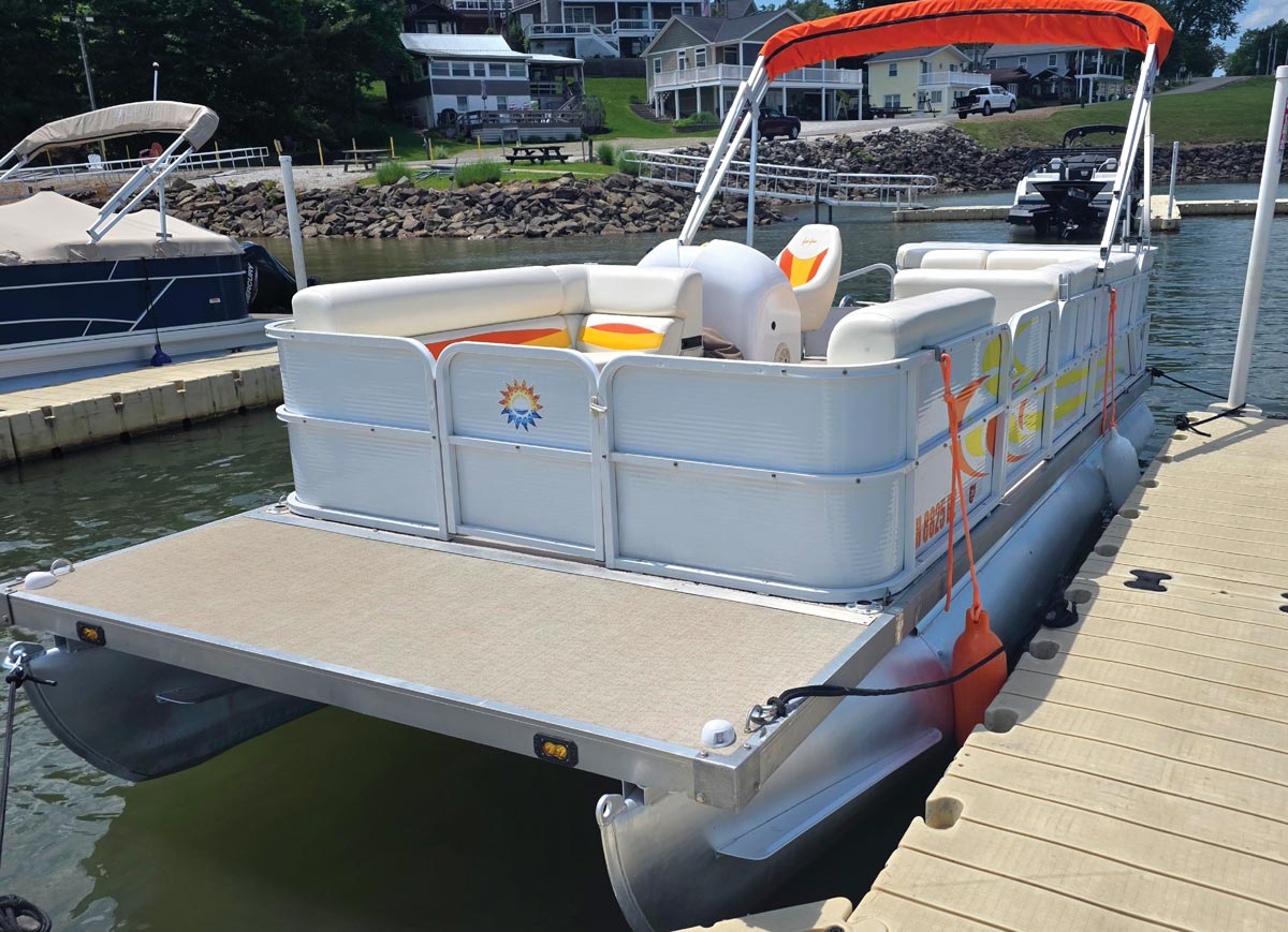 A white pontoon boat with custom orange decals and an orange bimini top docked at a floating pier.